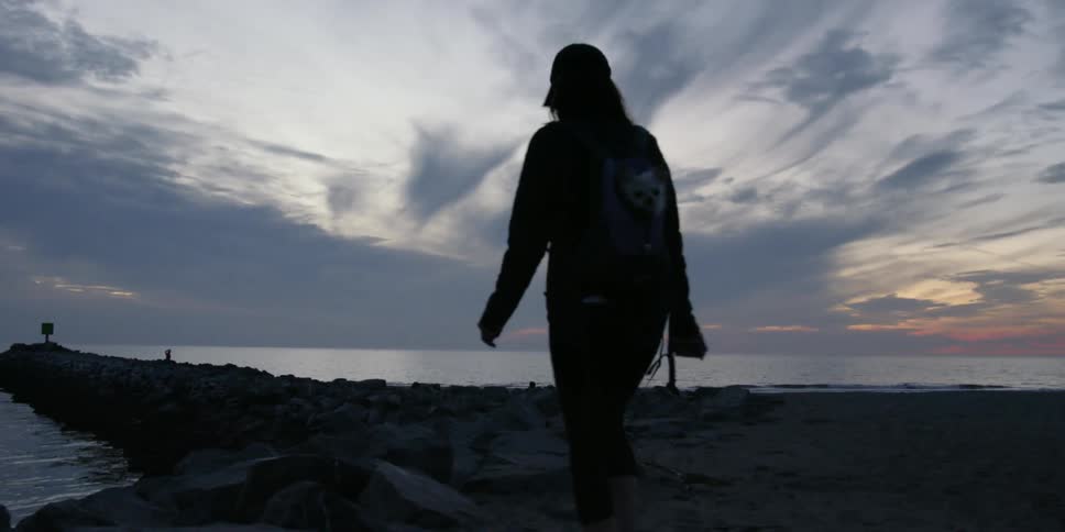 A woman carrying a dog in her backpack walks toward a rock jetty