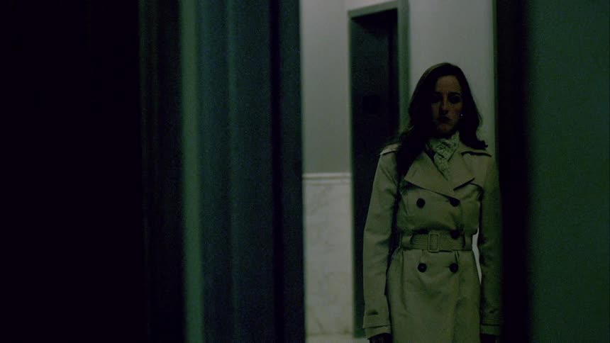A woman wearing a trench coat walks out of the elevator and down the hall