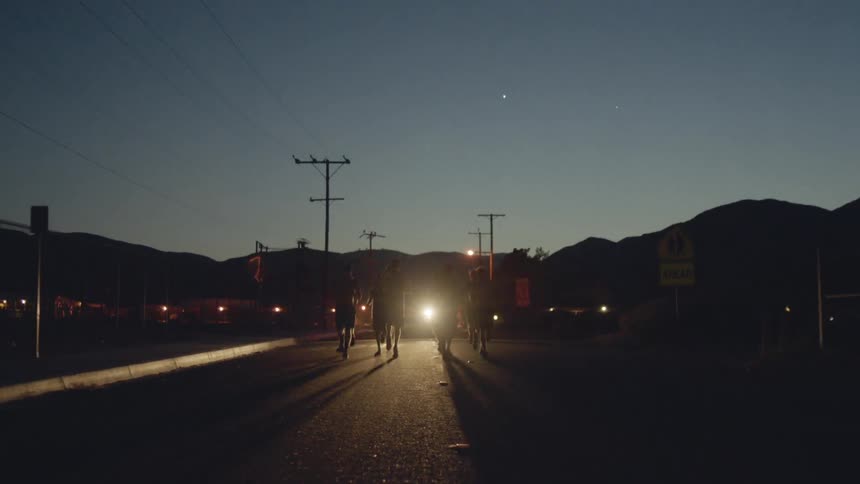 Group of people running down a road at sunset with only car headlights behind them