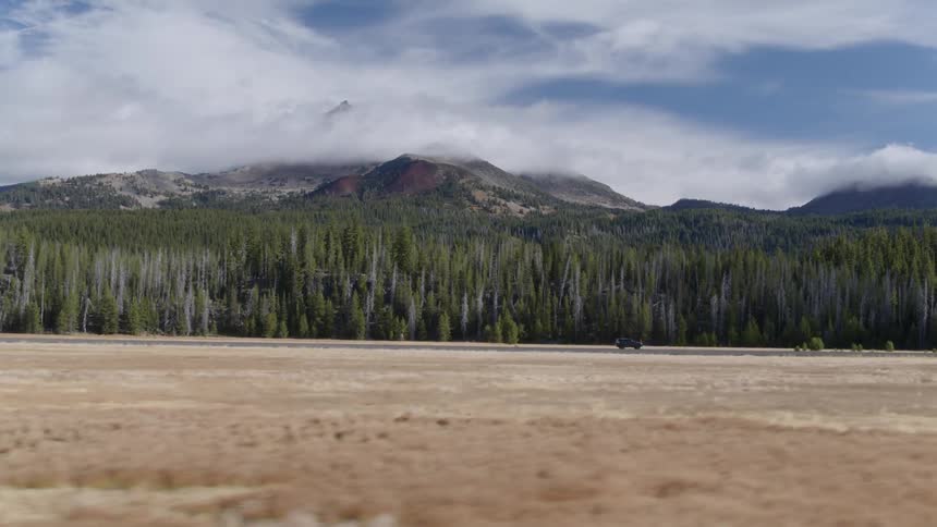 An suv is driving on a road past a forest and cloud covered mountain during the day