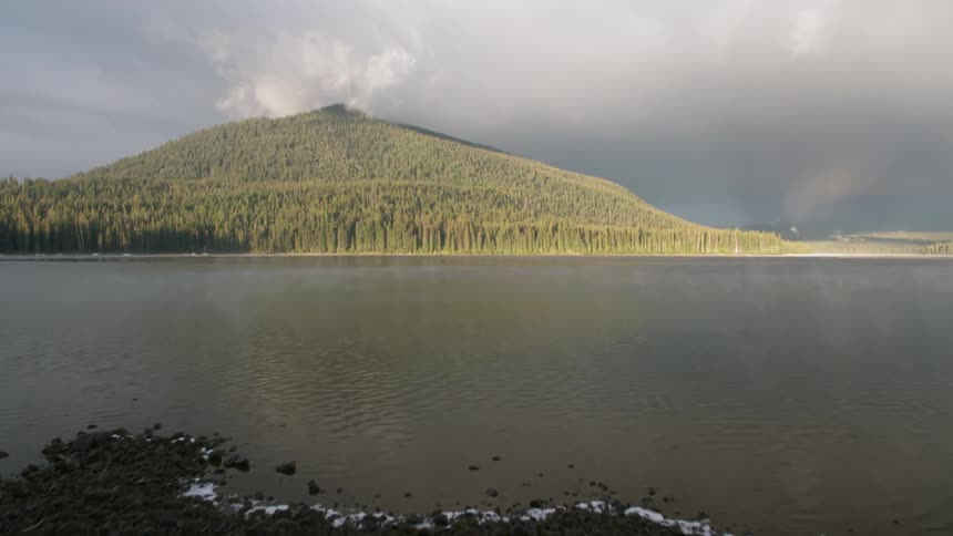 Storm clouds are hovering over a river and forest during the day