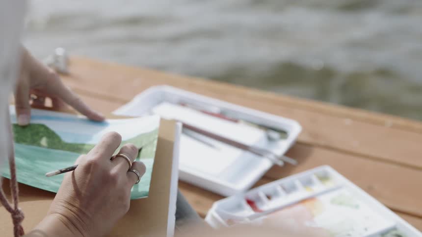 A woman is painting with watercolors as she sits on a dock during the day