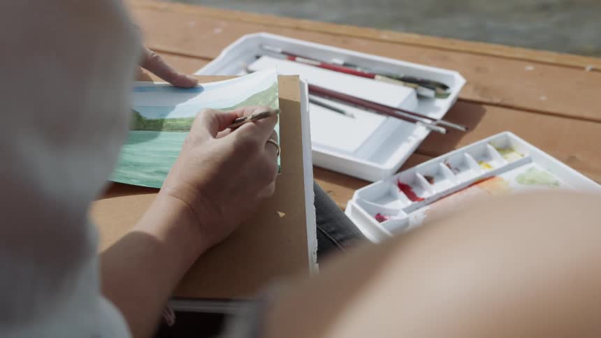 A woman is painting with watercolors as she sits on a dock during the day