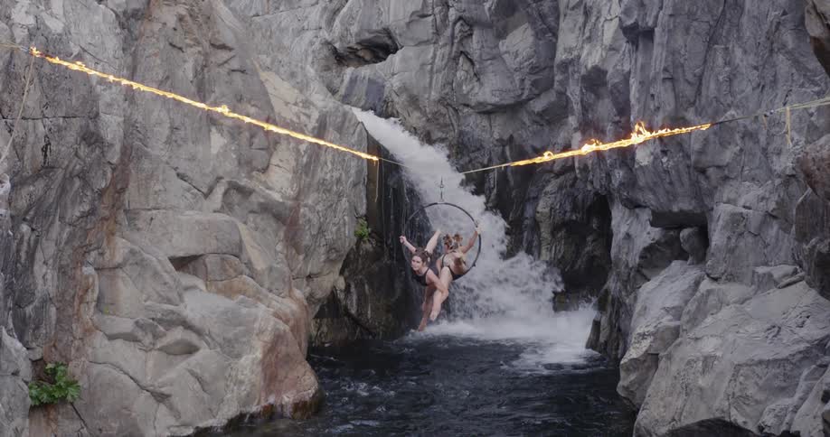 Two women twirl in a ring hanging from a flaming tightrope near a waterfall