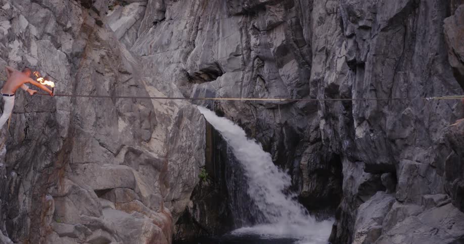 A man with a burning torch balances on a tightrope in a canyon