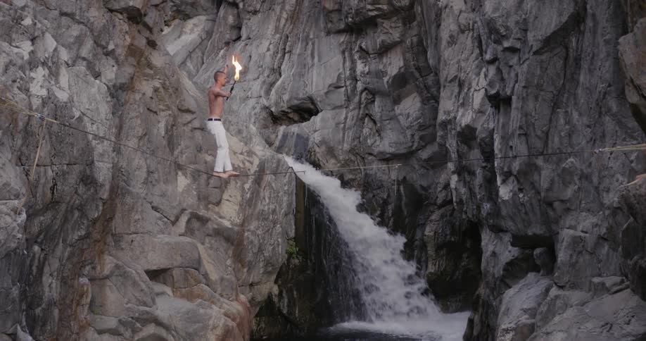 A man with a burning torch balances on a tightrope over a gorge with waterfall