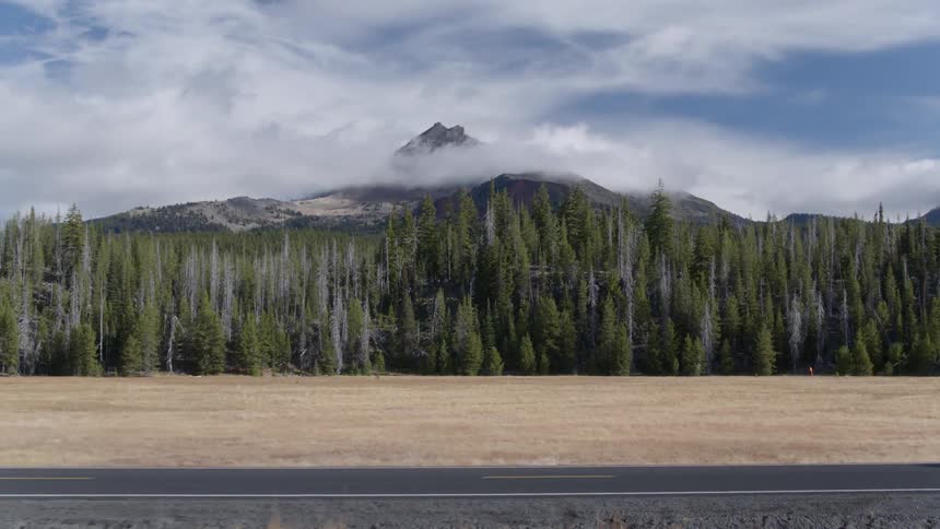 An suv is driving on a road past a forest and cloud covered mountain during the day