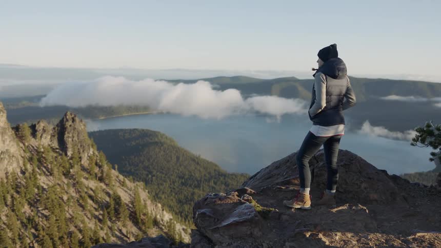 A woman stands on a ledge and looks down at the water and hills