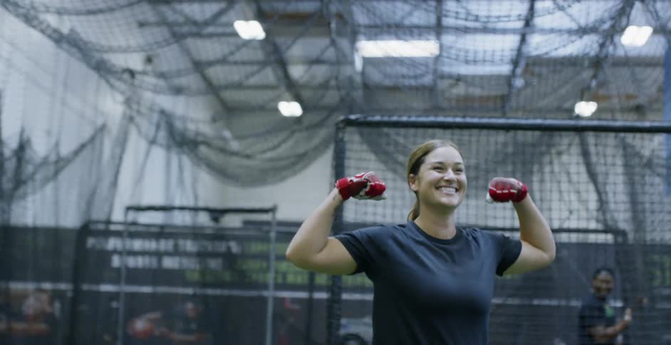 A female athlete is laughing and posing as she practices baseball indoors
