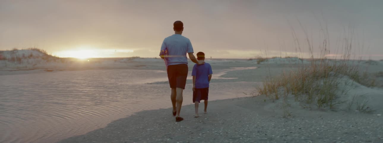 Man and boy walking on a beach carrying a kite at sunset
