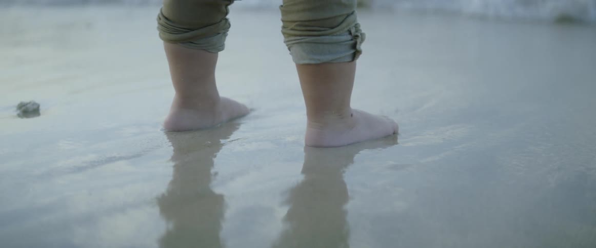 A child stands in sand with pants rolled up as waves come to shore