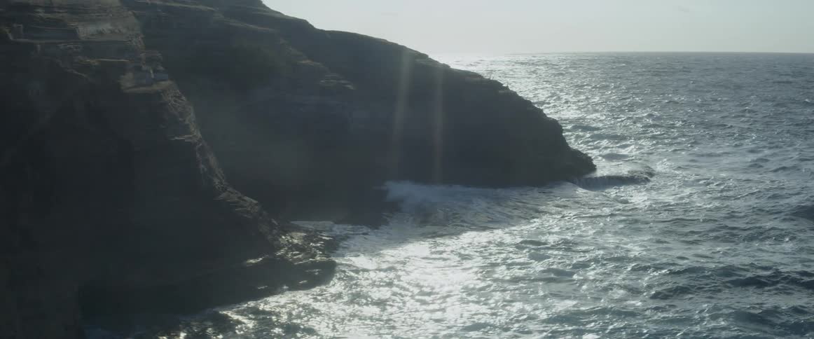 Ocean waves splash against the rock cliff shore on a sunny day