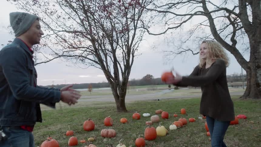 A man and woman throw a pumpkin back and forth on a farm