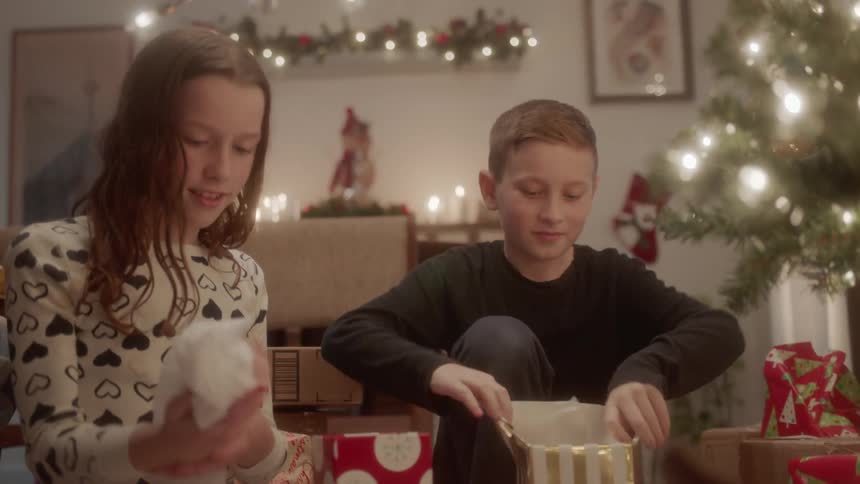 A girl and boy open presents next to a christmas tree