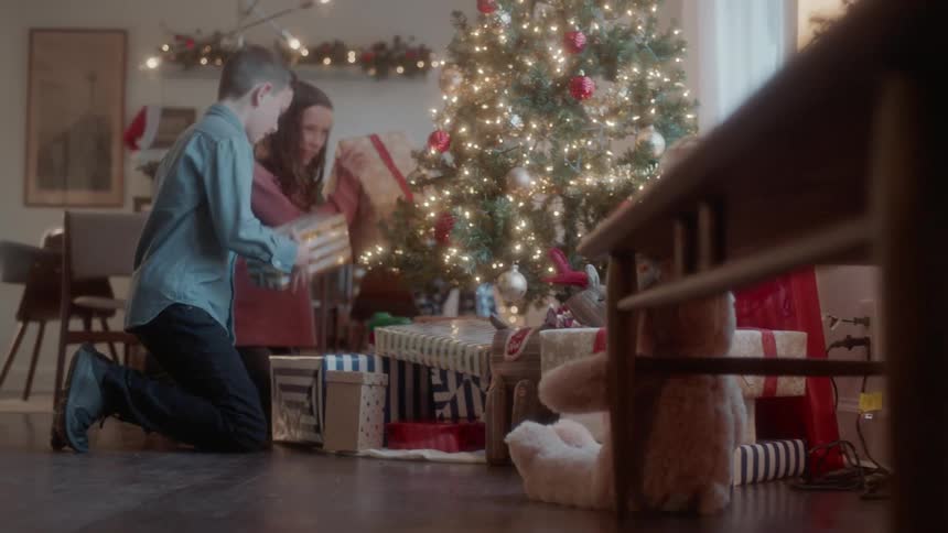 Children kneel down to shake christmas presents beneath a tree