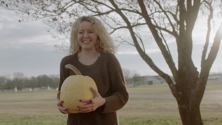 A woman holds a pale pumpkin by a tree