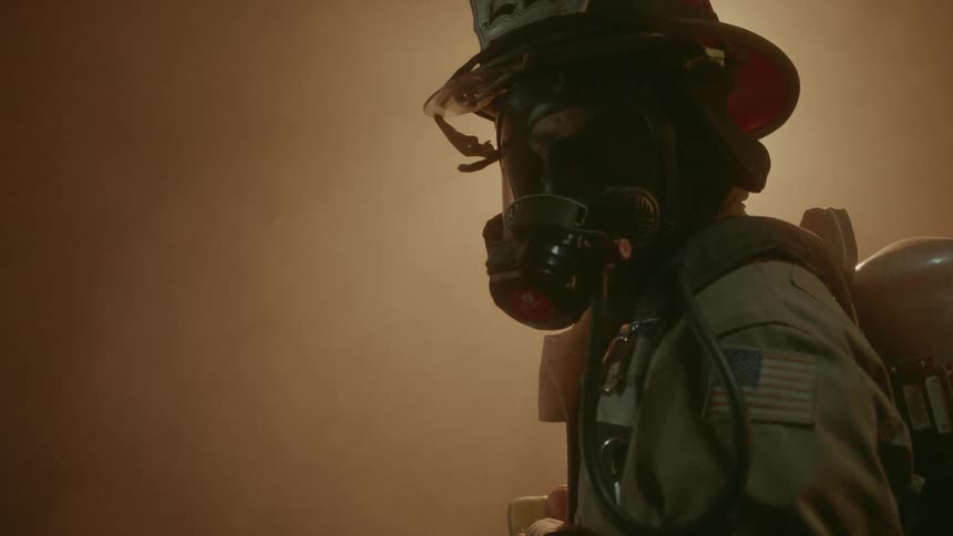 A firefighter wearing a mask and oxygen tank stands in front of a fire in the dark