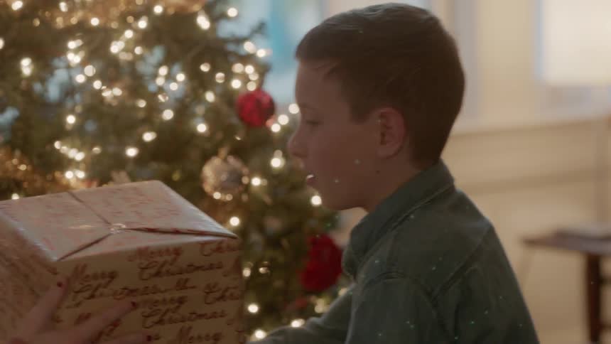 A girl and boy shake a present next to a christmas tree