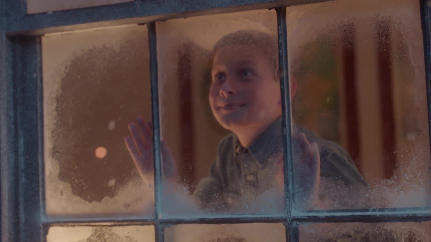 A kid is looking out the window at the snow falling