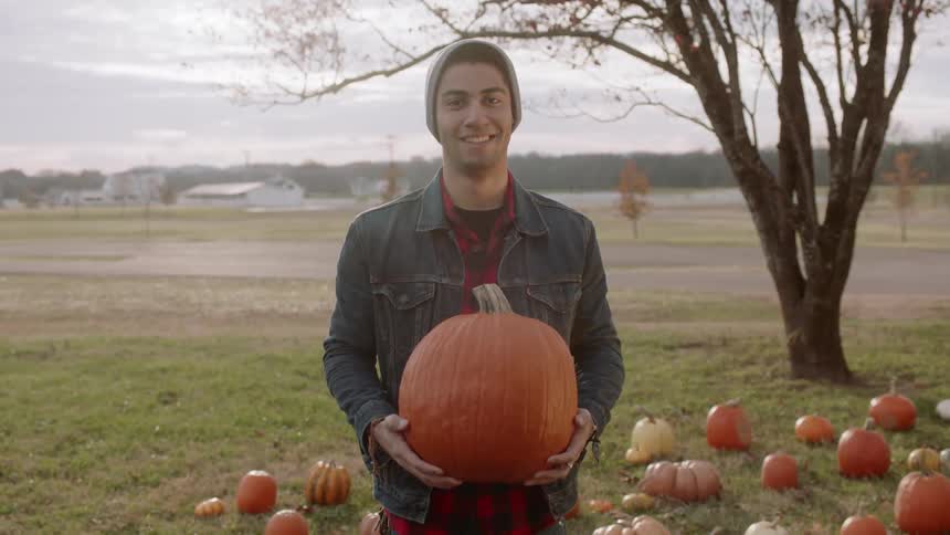 Man standing among pumpkins holds large pumpkin and smiles