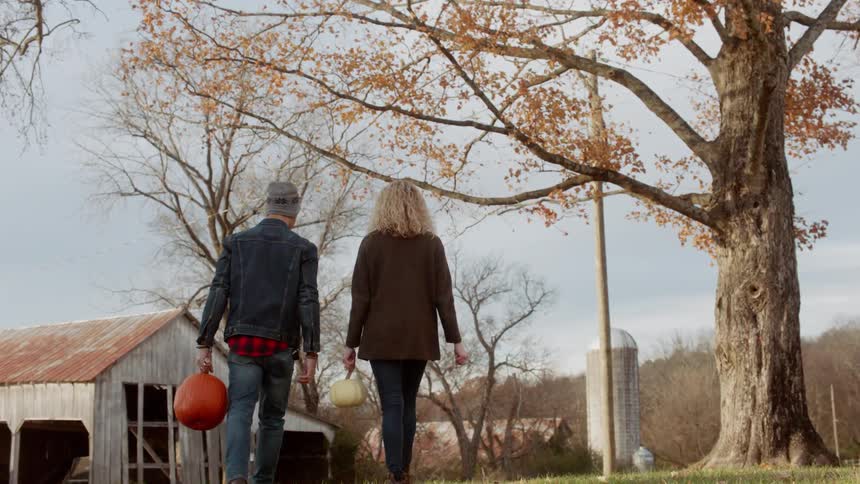 A couple carrying pumpkins walks toward a barn
