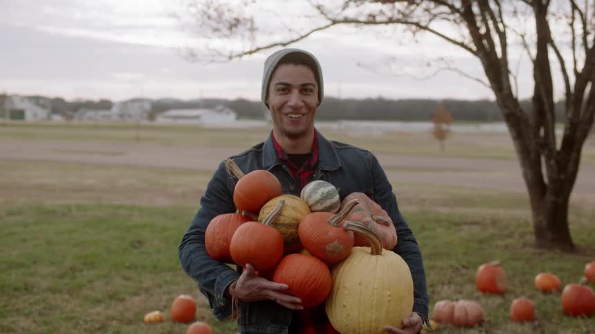 A man smiles as he is holding a bunch of pumpkins on a cloudy day