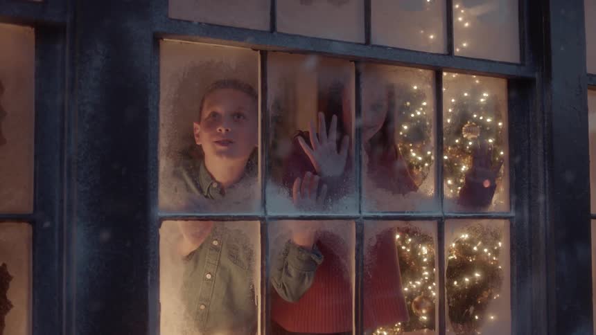 A boy and girl watch snow from a living room window on christmas