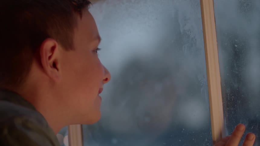 A boy is looking out of a window in his home watching snow fall