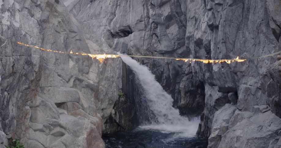 A man holding a torch does a backflip off a burning tightrope into the water