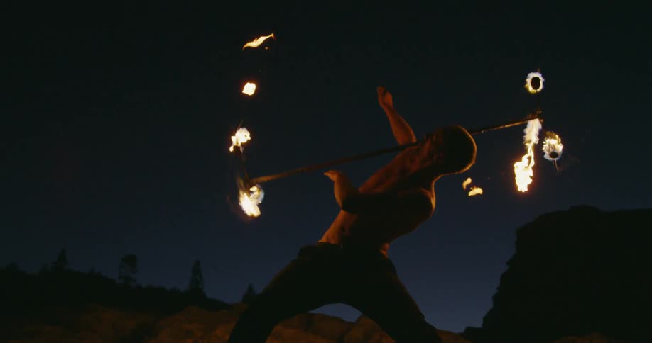A man twirls a rod with fire on each end in slow motion