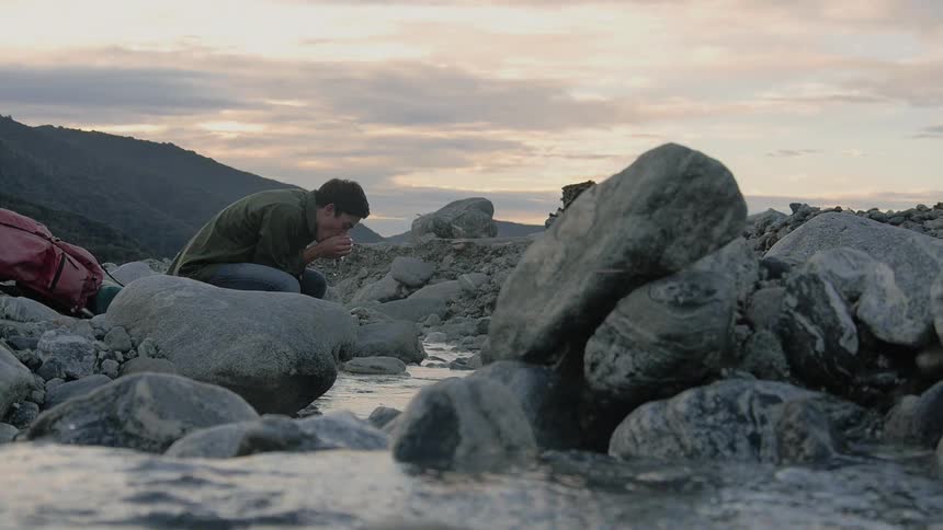 A young white male kneels by a stream and drinks water from his hands