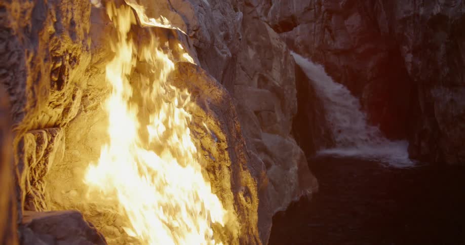 A fire is burning up a rock ledge near a waterfall at dusk
