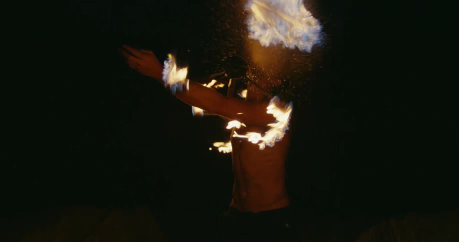A man is fire spinning and spits fire at camera