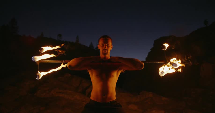 A serious tribal man holds fire burning staff at night and stares