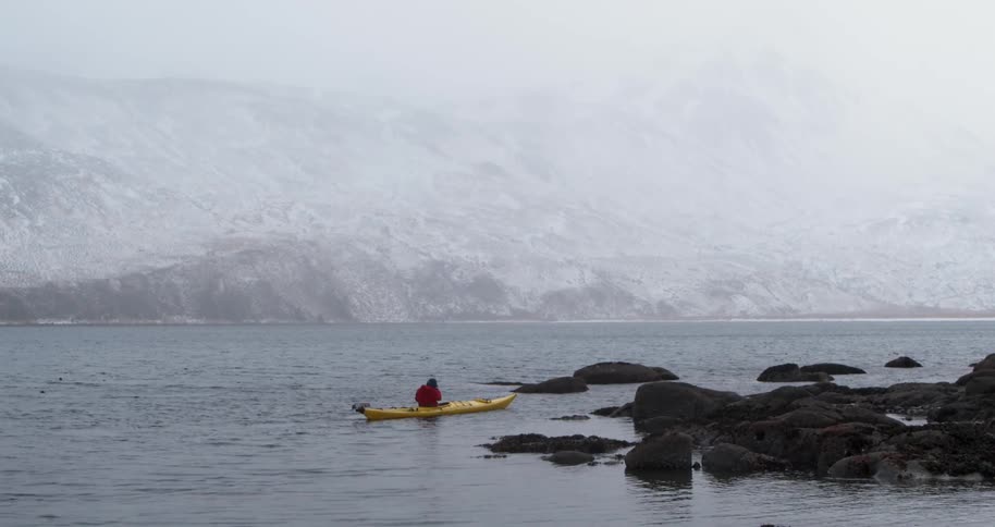 A man is paddling a kayak along a mountainous coastline in winter