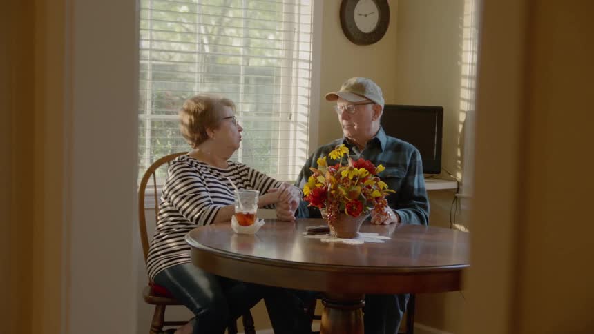 A husband and wife sit at a round kitchen table holding hands