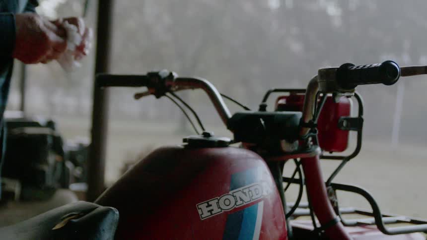 An older man is twisting the gas cap on an atv and getting on during the day