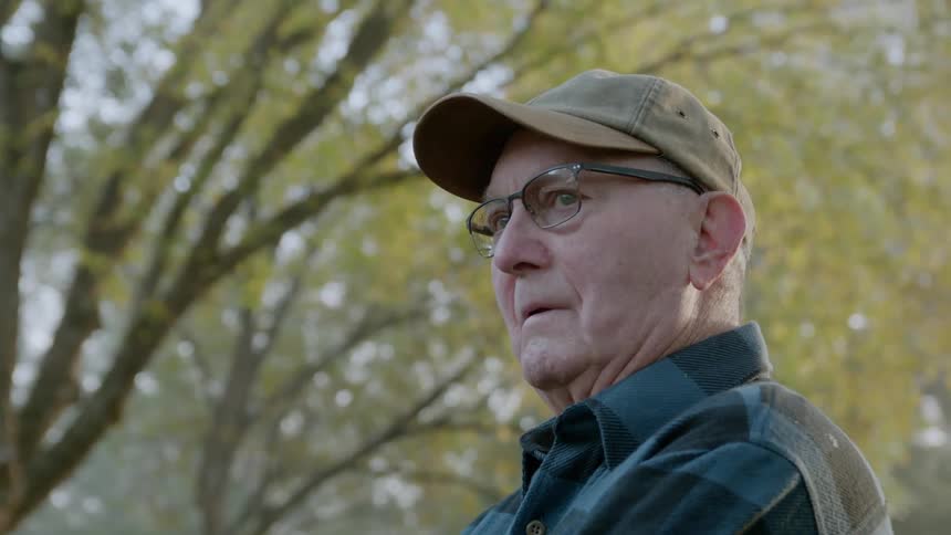 An older man is standing outside beneath trees looking around during the day