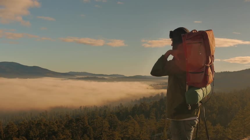 A man carries a backpack while looking over a forest from above