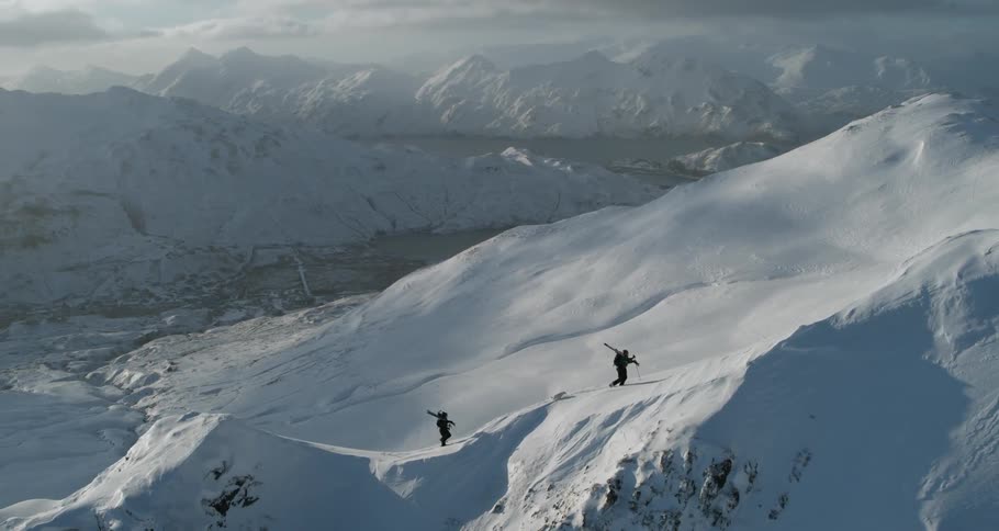 Two people carrying skis are climbing up a mountain ridge in winter