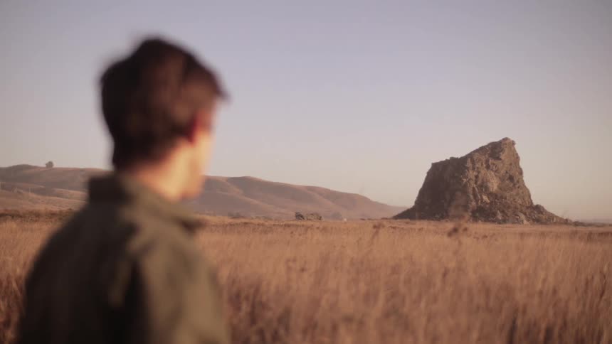 A young white man sits up in a field and then stands up and looks to a rock formation in the distance