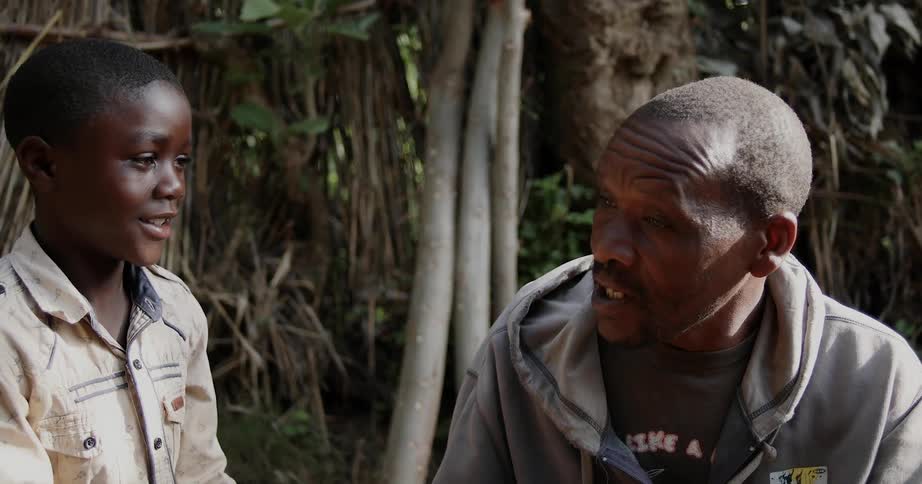 A boy is talking to his father outside near a hut in africa