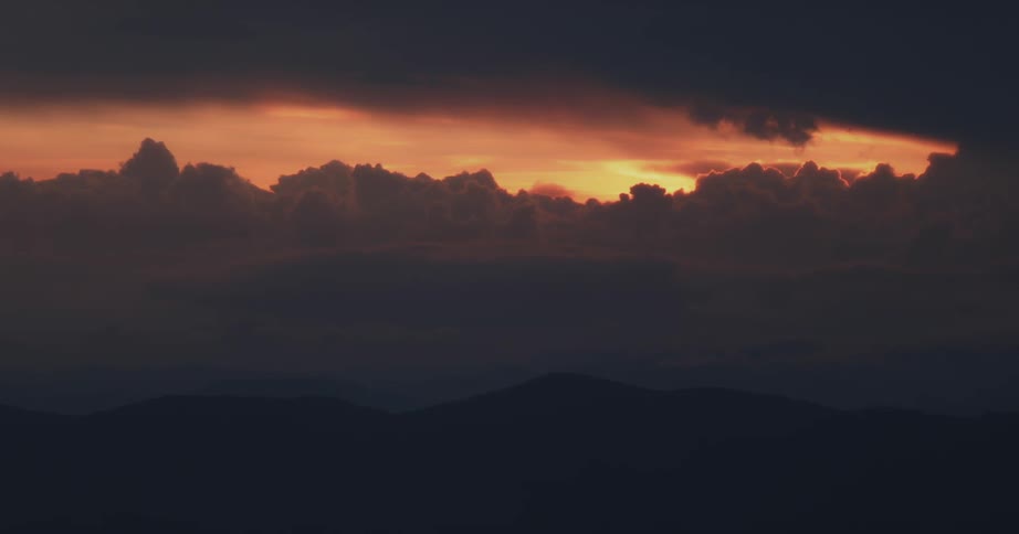 An orange sunset behind a line of mountains with dark clouds above
