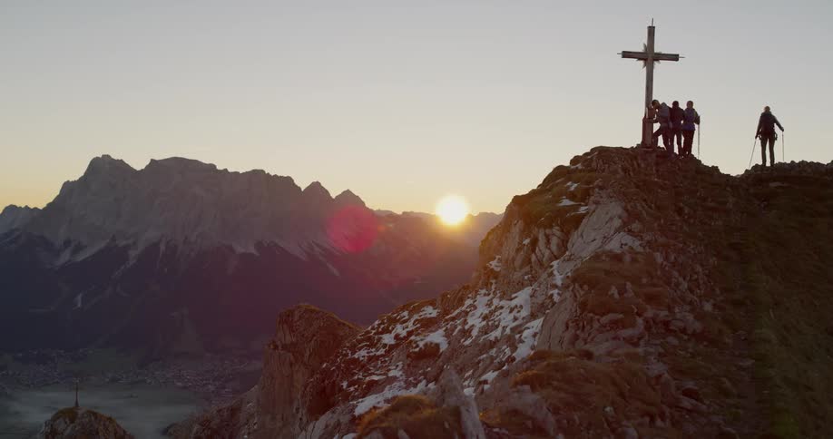 Hikers stand next to a cross on a mountain peak at sunset