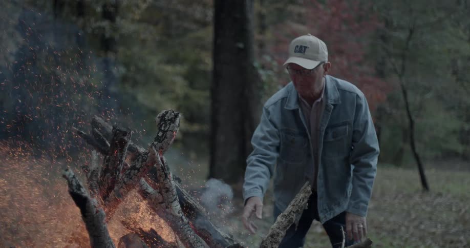 An old man is putting a log on a bonfire in a natural setting at dusk