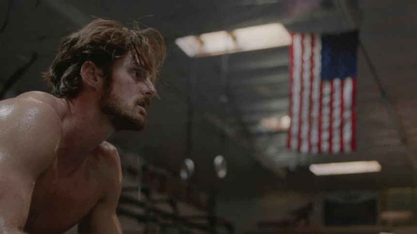 A man sits in a gym that has an american flag hanging from the rafters as he runs his hand through his hair