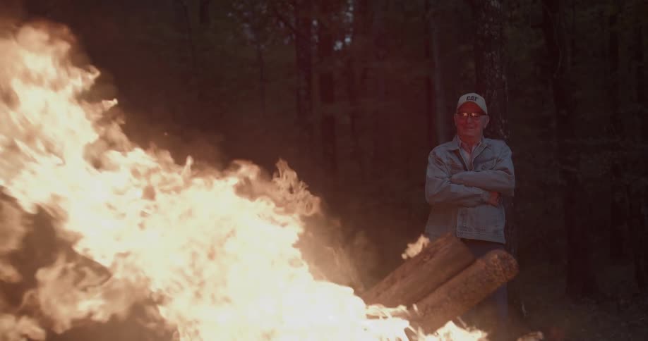 A man stands by a campfire in a wooded area at night with his arms crossed