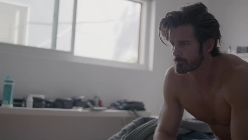 Man sitting on the end of a bed with hands clasped with a window open