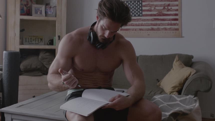 A shirtless man writes on a notebook while sitting on a coffee table