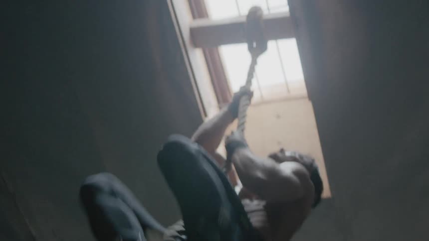 A male athlete is climbing up a rope in a gym during the day